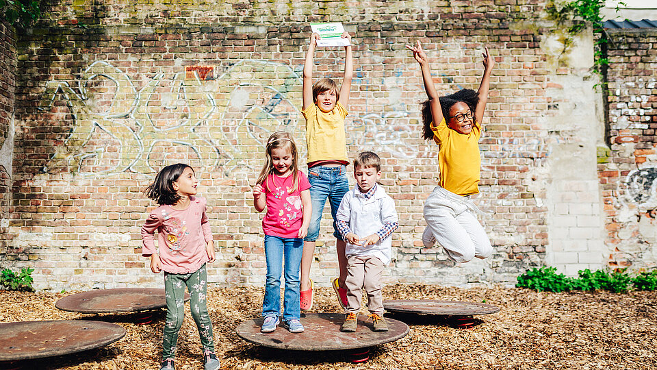 Fünf Kinder springen hoch und halten die Plakette hoch