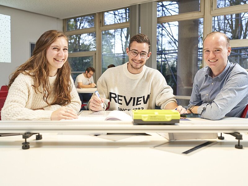 Eine Studentin, ein Student und eine Lehrperson nebeneinander sitzend an einem Gruppentisch