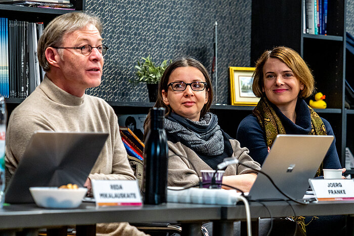 Das Bild zeigt einen Mann und zwei Frauen, die an Tischen sitzen.